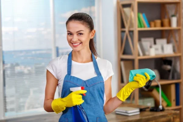 House cleaner in a blue apron holding a spray bottle and cloth in a bright living room, preparing a rental for the next guest in Chandler, AZ.