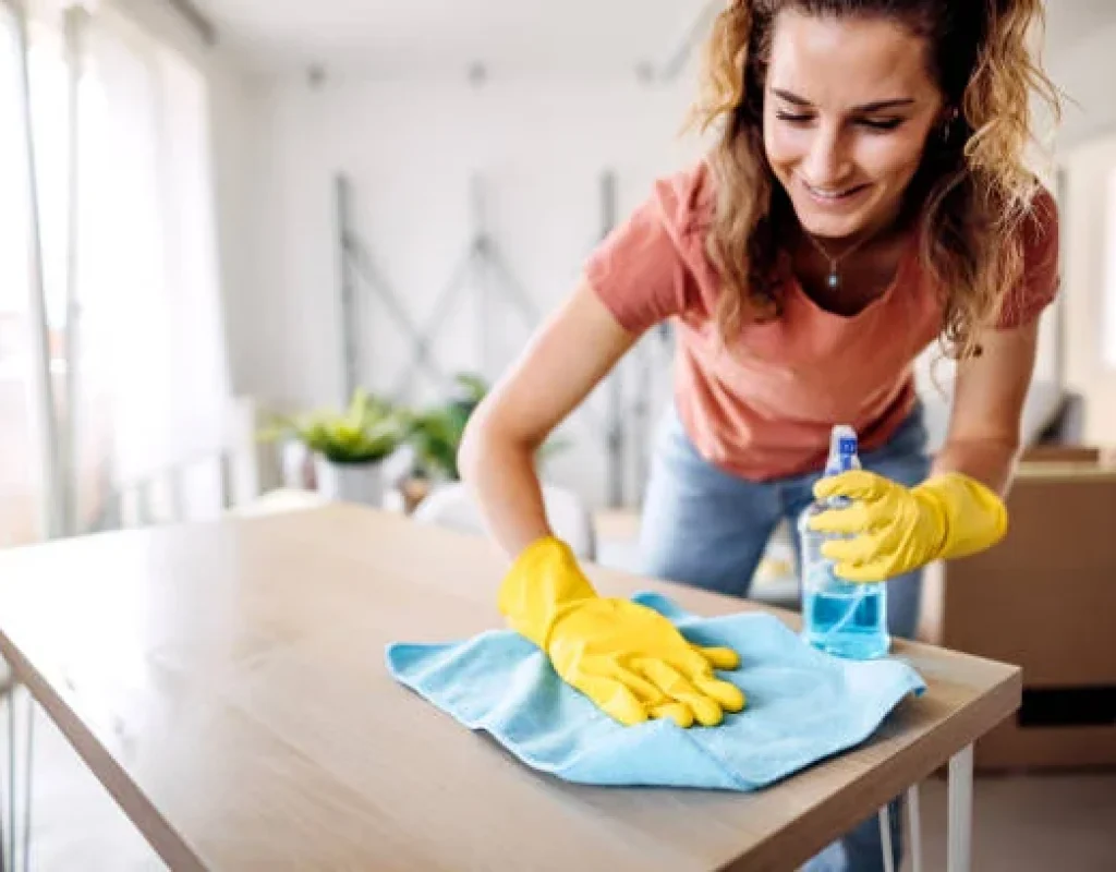 House cleaner in yellow gloves wiping down a wooden table with a blue cloth and spray bottle during a recurring cleaning visit in Scottsdale, AZ.